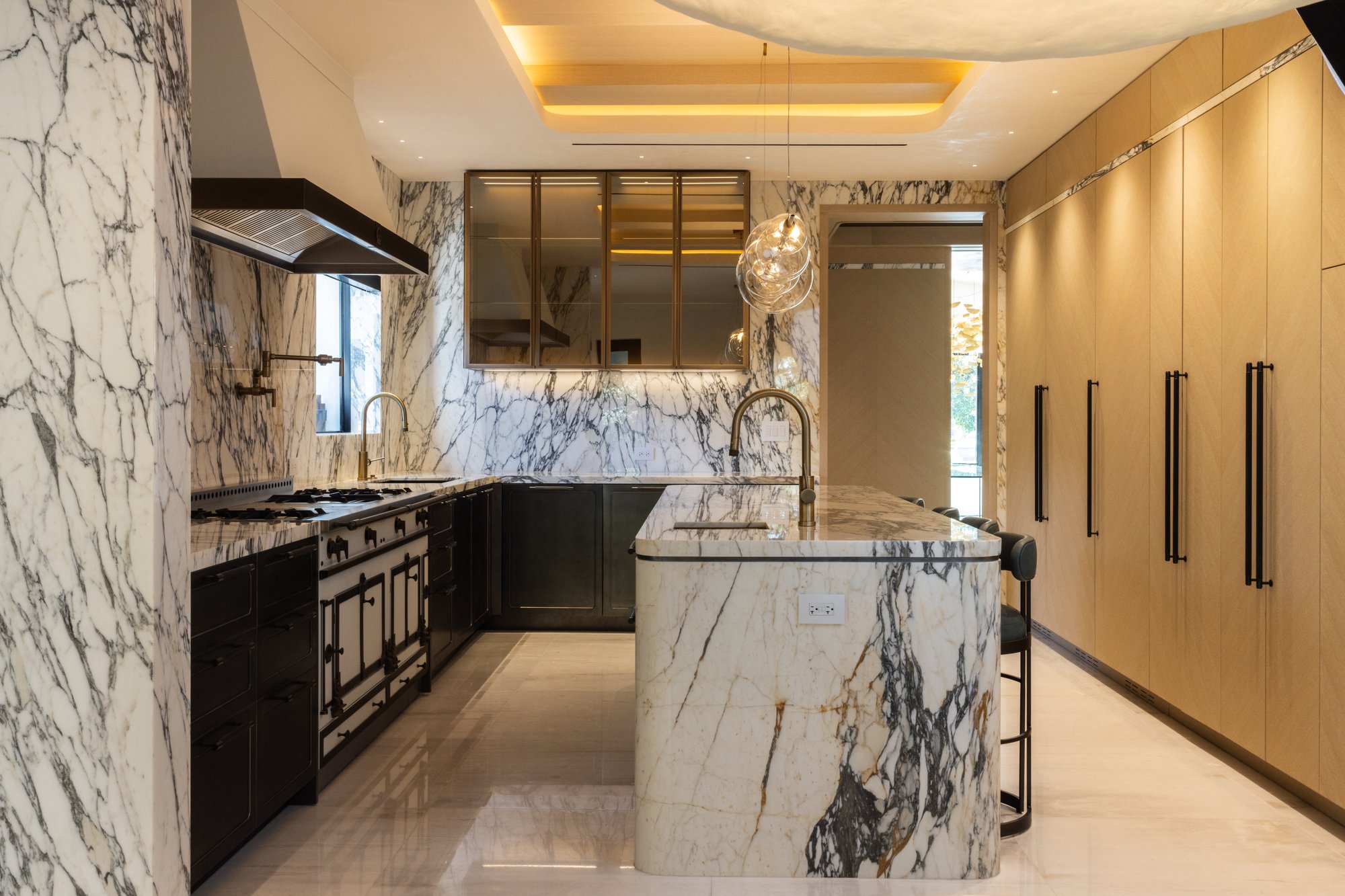 Luxury kitchen with Calacatta marble waterfall island and matching marble walls, a vintage-style black and gold range oven, and custom floor-to-ceiling light wood cabinetry.