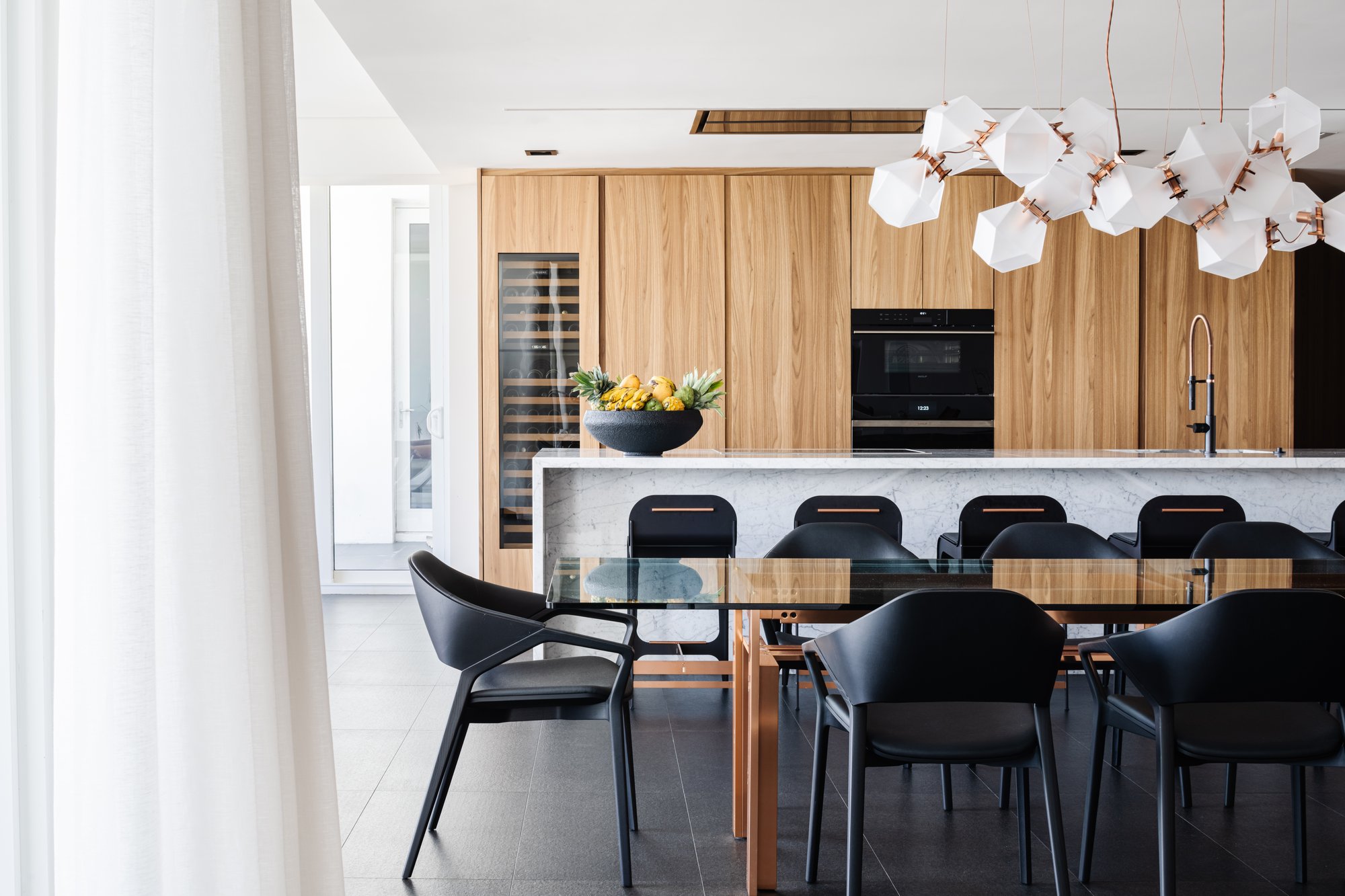 Modern open-concept kitchen and dining area featuring natural wood cabinetry, integrated wine storage, a white marble waterfall island, and a geometric designer chandelier.