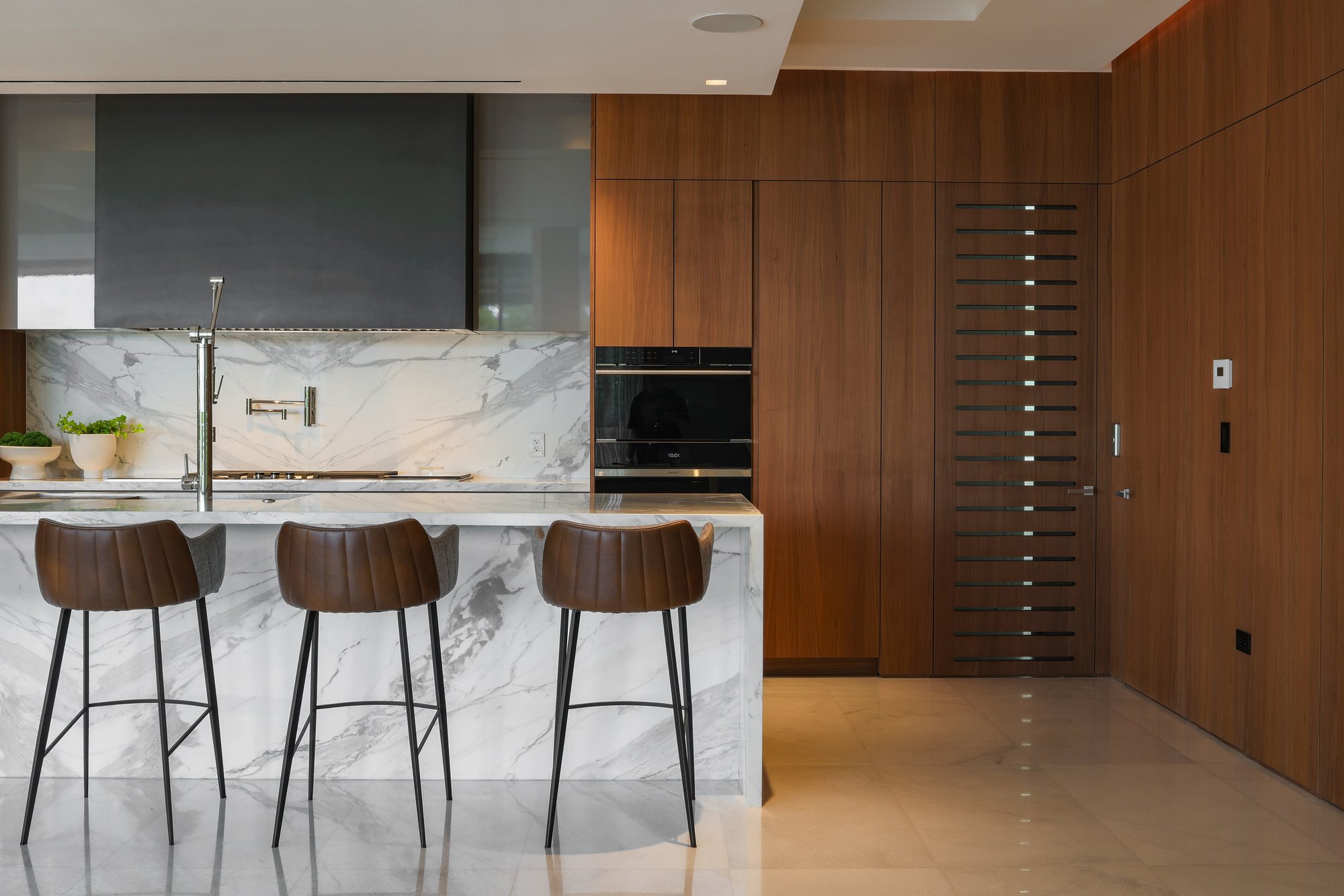Minimalist modern kitchen featuring floor-to-ceiling walnut wood cabinetry with integrated ovens, a full Calacatta marble backsplash, and a stunning marble waterfall island bar with three ribbed leather barstools.