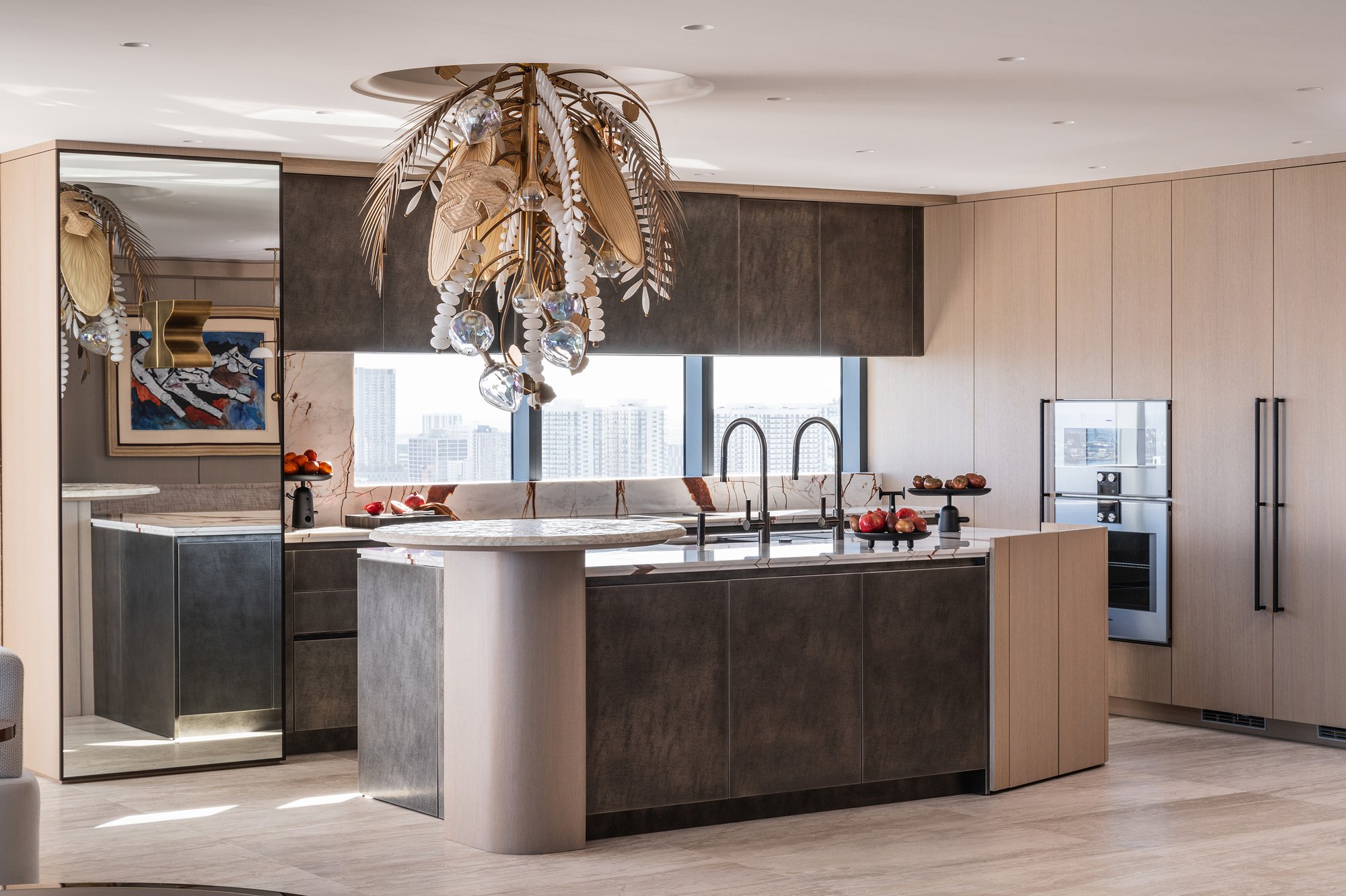 Luxury kitchen with a unique rounded travertine breakfast bar attached to a dark textured island, light wood custom cabinetry, and a large organic-style gold chandelier.