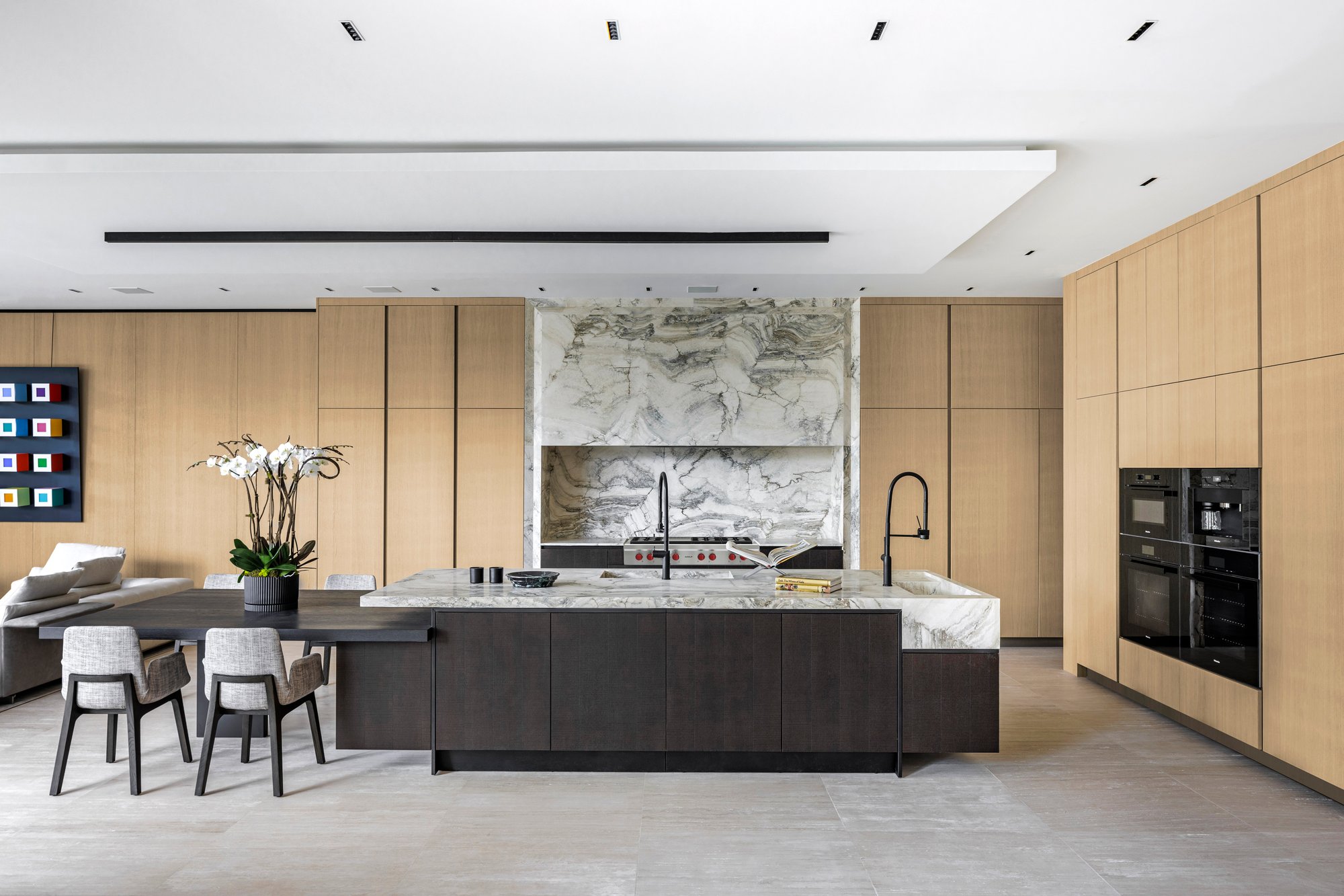 Modern open-concept kitchen with light wood cabinetry, a custom dark wood island, matching Calacatta marble counters and waterfall edges, and a dramatic bookmatched marble feature wall behind a Wolf range.