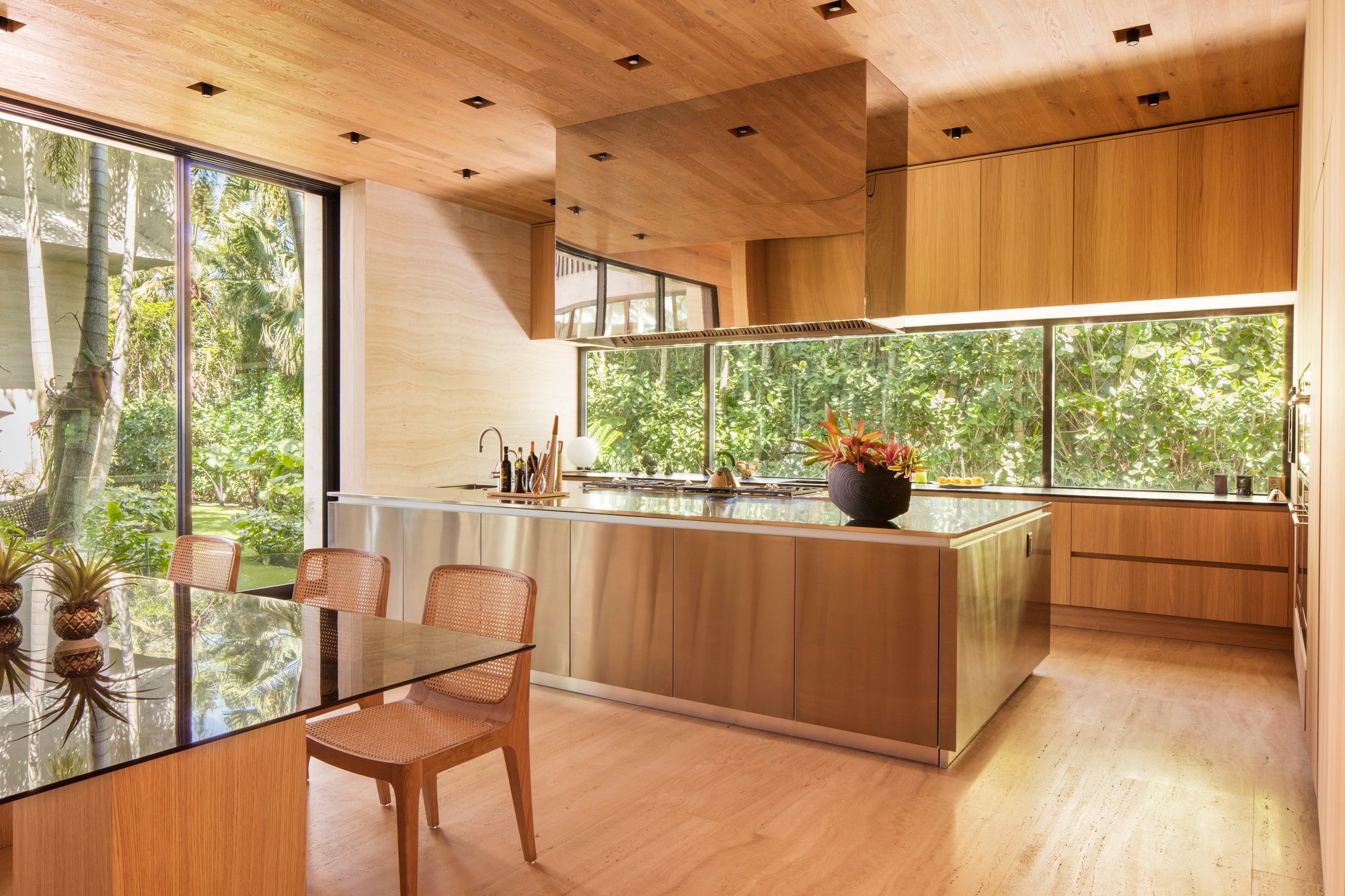 Tropical modern luxury kitchen featuring warm natural wood floors, walls, and ceilings, a massive brushed stainless steel island with an integrated cooktop, and a matching mirrored range hood.